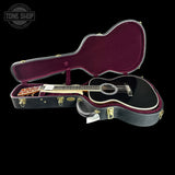 A black acoustic guitar inside an open black hard case with a burgundy velvet interior, shown on a solid black background. A Tone Shop watermark is visible in the top-left corner.