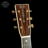 Close-up of the headstock of a Martin acoustic guitar against a solid black background, showing the dark wood grain, gold tuning pegs, and white 'MARTIN' lettering.