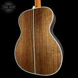 Close-up of the back of an acoustic guitar showing a rich brown wood grain finish and a white binding edge, set against a solid black background with a Tone Shop watermark in the top left.