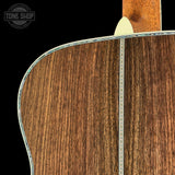 Close-up of the back of an acoustic guitar showing the rich brown wood grain of the soundboard, a silver metal tone ring, and part of the neck joint against a black background.