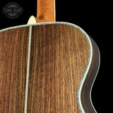 Close-up detail of an acoustic guitar's wooden back, showing the rich brown wood grain, a decorative inlay line, and part of the neck against a black background. A Tone Shop watermark is visible in the top left corner.