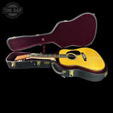 An acoustic guitar with a natural wood body and headstock, displayed inside an open black hard case with a burgundy interior, set against a solid black background. A 'TONE SHOP' logo is visible in the top-left corner.