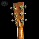 Close-up of the headstock of an acoustic guitar with a glossy brown wood finish and ornate gold tuning pegs, shown against a solid black background.