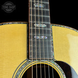 Close-up of an acoustic guitar body and neck, showing a natural wood grain top, dark fretboard, and iridescent mother-of-pearl inlays against a black background. A Tone Shop watermark is visible in the top left corner.