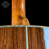 Close-up detail of an acoustic guitar body and neck joint, showing rich wood grain, a decorative inlay line, and part of the glossy neck against a black background. A Tone Shop watermark is visible in the upper left.