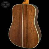 Close-up of the back of an acoustic guitar showing a rich brown wood grain finish with a light-colored center stripe and binding, set against a solid black background.
