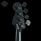 Close-up of the headstock of a black bass guitar, showing the four tuning pegs and tuning mechanism against a black background, with a Tone Shop watermark in the top left corner.