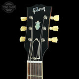 Close-up of a Gibson acoustic guitar headstock with six cream-colored tuning pegs, silver tuning keys, and a black glossy finish against a black background.