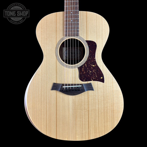 A Taylor acoustic guitar with a natural flamed maple top, dark brown pickguard, and black bridge, shown against a solid black background with a Tone Shop watermark in the top left corner.