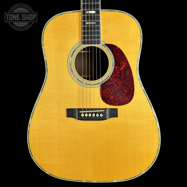 A front-facing acoustic guitar with a natural wood top, dark fretboard, and red pickguard, shown against a black background with a Tone Shop watermark in the upper left.