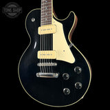 A black electric guitar with a cream pickguard and cream humbucker pickups, shown against a solid black background. A Tone Shop watermark is visible in the top left corner.