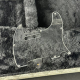 A clear plastic guitar pickguard with pre-drilled screw holes, shown resting on a dark gray faux fur surface.