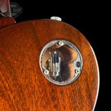 Close-up of the back of a vintage electric guitar body, showing a circular chrome hardware plate with a clear cover over a gold-toned pickup, on a black background.