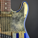 Close-up of a vintage-style electric guitar body with a heavily distressed silver pickguard, gold hardware, and a blue glitter-finish edge, set against a dark gray background.