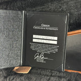 A close-up of a Gibson Certificate of Authenticity document inside an open black presentation case, with a small leather Gibson tag resting in front on a dark textured surface.