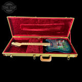 An electric guitar with a blue and green flame finish sits inside an open gold hard case with a red velvet lining, shown against a black background with a Tone Shop watermark in the top left corner.
