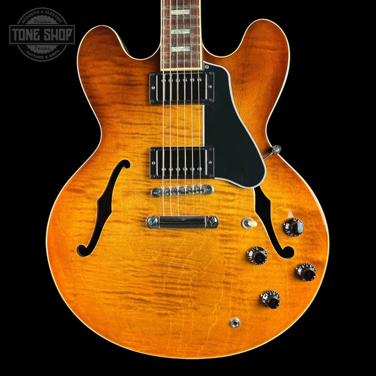 An electric guitar with a vibrant orange sunburst finish, black pickguard, and chrome hardware, shown against a solid black background with a Tone Shop watermark in the top left corner.