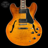 An electric guitar with a vibrant orange sunburst finish, black pickguard, and chrome hardware, shown against a solid black background with a Tone Shop watermark in the top left corner.