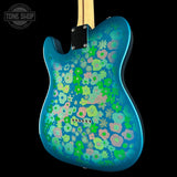 A close-up of the back of an electric guitar body featuring a vibrant blue metallic finish covered in a dense pattern of retro-style flowers in green, pink, and light blue. The neck is light wood, and a black plate is visible at the top, set against a solid black background.