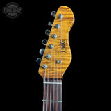 Close-up of an electric guitar headstock with a natural wood finish, six tuning pegs, and the 'TMC' logo, shown against a solid black background.