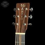 Close-up of the headstock of a Martin acoustic guitar against a black background, showing the brand name 'MARTIN' in gold on a dark wood headstock with six silver tuning pegs.