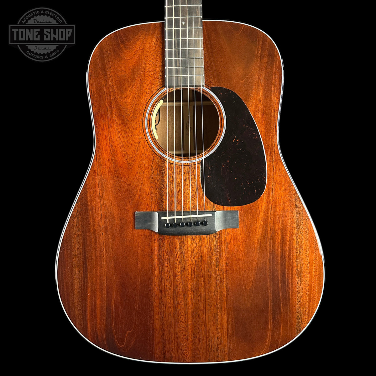 A close-up studio shot of an acoustic guitar body showing a rich, warm brown wood grain finish, a black pickguard, and a silver bridge, set against a solid black background with a Tone Shop watermark in the top-left corner.