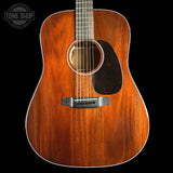 A close-up studio shot of an acoustic guitar body showing a rich, warm brown wood grain finish, a black pickguard, and a silver bridge, set against a solid black background with a Tone Shop watermark in the top-left corner.