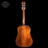 The back of an acoustic guitar with a rich, warm brown wood finish and silver tuning keys, shown against a solid black background with a Tone Shop watermark in the top-left corner.