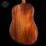 Close-up of the back of an acoustic guitar showing a rich, warm brown wood grain finish, set against a solid black background with a Tone Shop watermark in the top left corner.