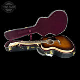 An acoustic guitar with a sunburst finish displayed inside an open black hard case with a burgundy interior, set against a solid black background. A Tone Shop watermark is visible in the top-left corner.
