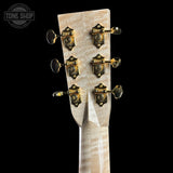 Close-up of the headstock of a guitar with a light natural wood finish and six ornate gold-toned tuning pegs, shown against a solid black background.