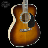 A close-up of an acoustic guitar body showing a classic sunburst finish that transitions from a bright amber-orange center to dark brown and black edges. The light natural wood neck and headstock are visible on the left side, set against a solid black background with a 'TONE SHOP' watermark in the upper left corner.