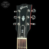 Close-up of a Gibson acoustic guitar headstock with six tuning pegs, showing the Gibson logo, a silver hand-painted bird emblem, and 'SC' on the black glossy finish against a black background.