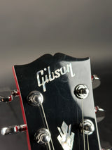 Close-up of a Gibson electric guitar headstock, showing the Gibson logo in silver script on a glossy black background with silver tuning pegs and strings.
