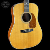 An acoustic guitar with a natural wood finish and a dark brown pickguard, shown against a solid black background with a Tone Shop watermark in the top left corner.
