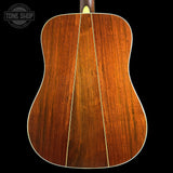 Close-up of the back of an acoustic guitar showing a rich, warm brown wood grain finish with a double-cutaway body shape and a white binding edge, set against a solid black background.