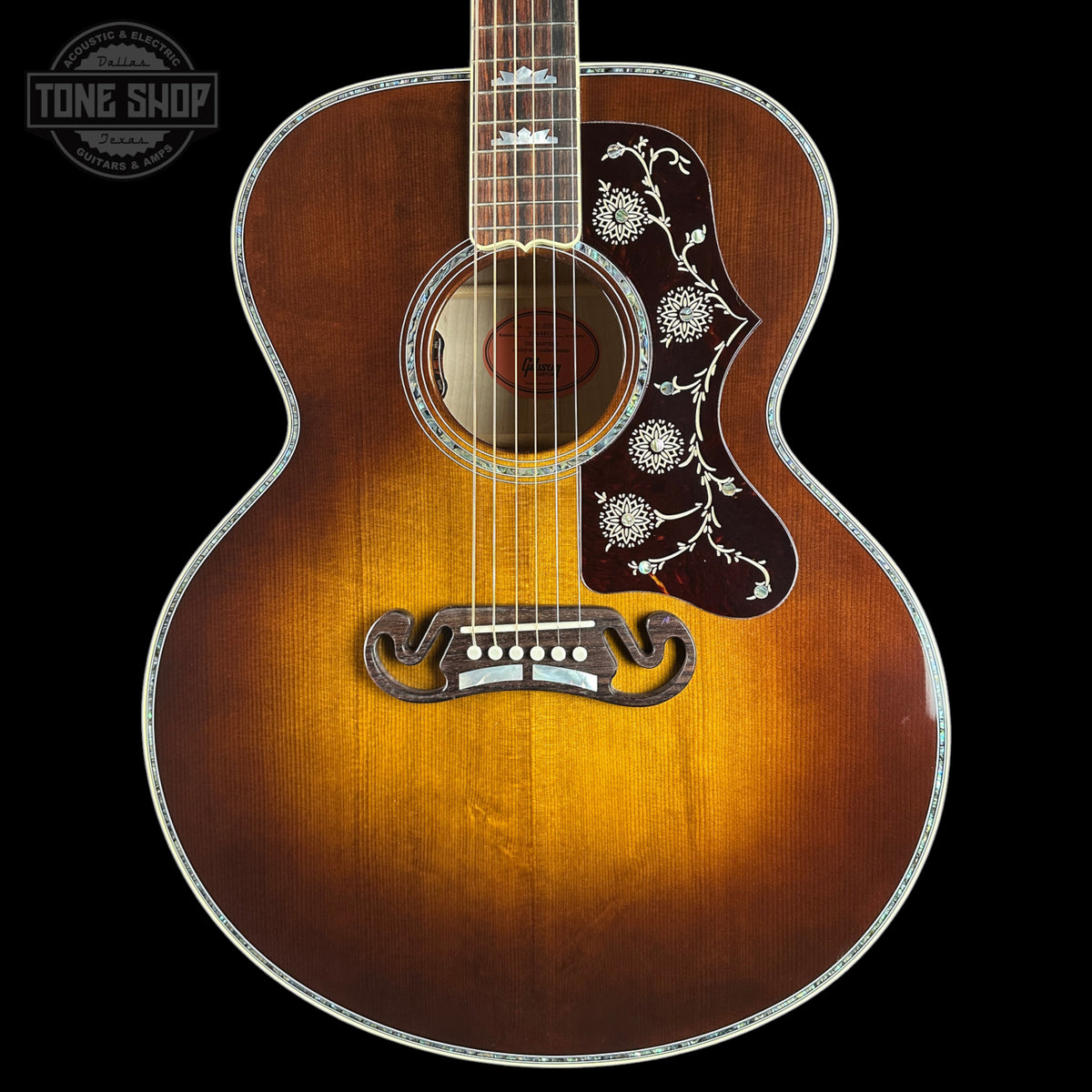 A vintage-style acoustic guitar with a sunburst finish, featuring floral inlay on the pickguard and a decorative trapezoid soundhole rosette, shown against a solid black background.