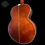 Close-up of the back of an acoustic guitar showing a rich reddish-brown wood finish with a light-colored center stripe and white binding, set against a solid black background. A Tone Shop watermark is visible in the top-left corner.