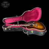 An acoustic guitar with a sunburst finish resting inside an open black hard case with bright pink velvet lining, shown against a solid black background. A Tone Shop watermark is visible in the top-left corner.