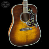 A vintage-style acoustic guitar with a natural sunburst finish, featuring a decorative pickguard with white and yellow bird and butterfly motifs, shown against a solid black background.