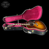 An acoustic guitar with a sunburst finish displayed inside an open black hard case with a magenta-pink velvet interior, set against a solid black background. A Tone Shop logo is visible in the top-left corner.