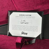 A Gibson Custom Certificate of Authenticity card inside a black frame, placed on a deep magenta velvet surface with a black strap visible at the side.