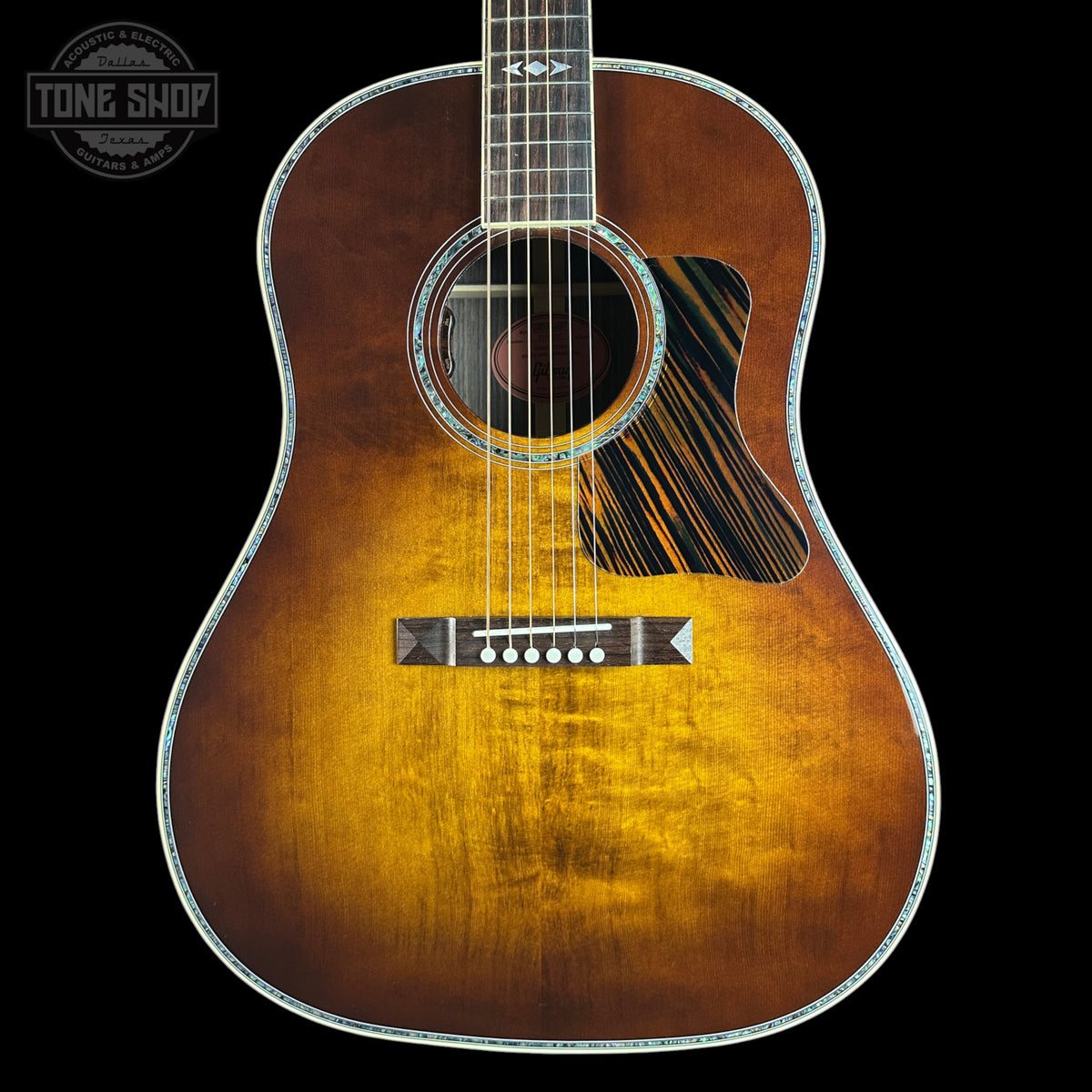 A front-facing acoustic guitar with a natural sunburst finish, featuring a dark pickguard and a silver-toned metal truss rod cover, set against a solid black background.