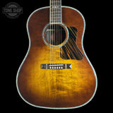 A front-facing acoustic guitar with a natural sunburst finish, featuring a dark pickguard and a silver-toned metal truss rod cover, set against a solid black background.