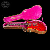A red electric guitar inside an open vintage brown hard case with a bright pink velvet interior, shown against a solid black background. A Tone Shop watermark is visible in the top-left corner.