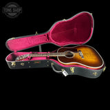 An acoustic guitar with a sunburst finish displayed inside an open hard case with a bright pink interior, set against a solid black background. A 'TONE SHOP' watermark is visible in the top-left corner.