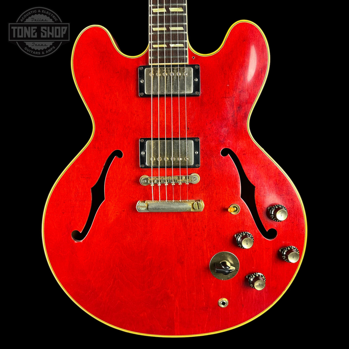 A red electric guitar with a glossy cherry finish, featuring black f-holes, dual humbucker pickups, and gold-toned hardware, shown against a solid black background with a Tone Shop watermark in the top left corner.