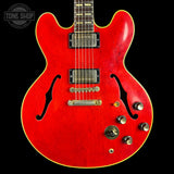 A red electric guitar with a glossy cherry finish, featuring black f-holes, dual humbucker pickups, and gold-toned hardware, shown against a solid black background with a Tone Shop watermark in the top left corner.