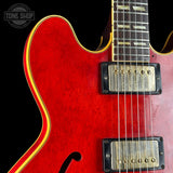 Close-up of a red electric guitar body with gold and yellow binding, dual humbucker pickups, and part of the dark fretboard against a black background. A Tone Shop watermark is visible in the top-left corner.