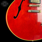 Close-up of a red electric guitar body with gold hardware, a black f-hole cutout, and a black background. A Tone Shop watermark is visible in the top left corner.